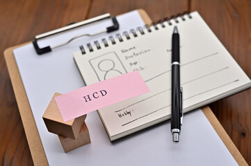 A note with the user's persona written on it is placed on a clipboard along with a pen. A sticky note stamped with "HCD" is placed on a wooden cube.