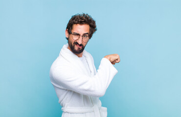 young bearded man wearing a bath robe feeling happy, satisfied and powerful, flexing fit and...