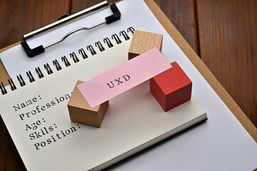 A note with the user's persona written on it is placed on a clipboard. A sticky note stamped with "UXD" is placed on a wooden cube.