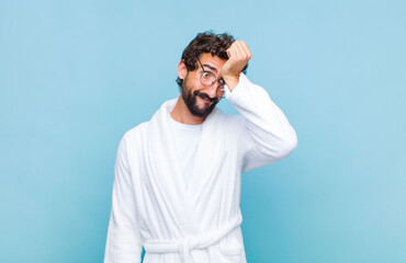 young bearded man wearing a bath robe raising palm to forehead thinking oops, after making a stupid...