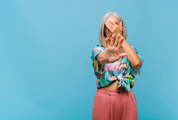 middle age cool woman covering face with hand and putting other hand up front to stop camera,...
