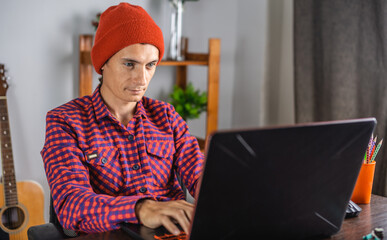 Fototapeta premium Modern young man in a red checked shirt and hat is working on a laptop. Concept of distance communication and remote job