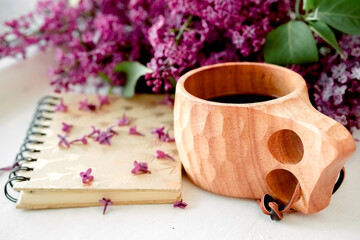 Wooden finnish cup with coffee on the table. Nearby lies a closed notebook for notes and a bouquet of lilacs. Good morning and good mood, planning a day at breakfast, concept.