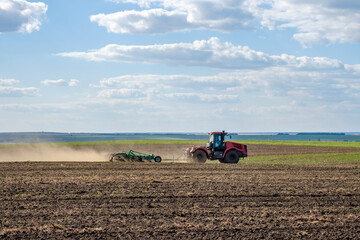 Obraz premium A red farm tractor in a cloud of dust cultivates the soil in the field with a cultivator after harvest. Summer sunny day. Fertile land. Modern agricultural machinery. Copy space. High quality photo