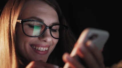 Beautiful young Woman Lying in Bed, Using Smart Phone and Smiling before Sleep. Woman using Cellphone on bed at Night. Attractive Girl looking at Mobile Phone.