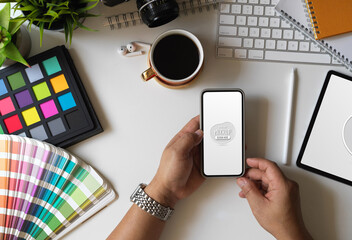 Designer holding mock up smartphone in his workspace with Pantone swatches and office accessories