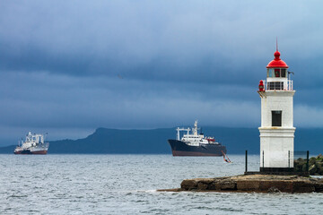 Fototapeta na wymiar lighthouse on Cape Tokarevskaya cat in Vladivostok against the background of ships on the roadstead in cloudy weather