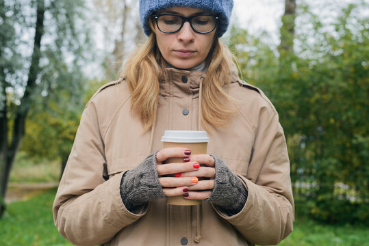 caucasian young woman in autumnal casual clothes standing alone, looking down, holding in hands paper cup of coffee on park trees background, horizontal outdoors lifestyle stock photo image