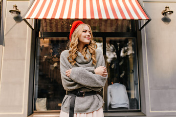 A cozy photo of a cute, charming blonde with curly hair, tenderly and mysteriously looking into the distance against on the background of a beautiful building facade.
