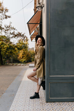 Stunning, charming blonde in a beige trench coat standing on the street, leaning on the house. Full-length photo against a gray building.