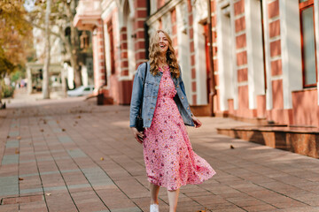 Naklejka premium smiling curly blonde goes down the street in a denim jacket and a light pink full-length sundress against the background of the building