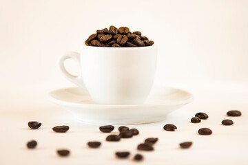 Almost white still life with a small white cup full of dark roasted coffee beans on a white saucer on a white background. Some beans are scattered in the foreground out of focus