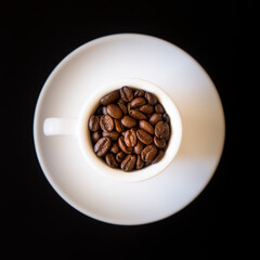 Still life with dark roasted coffee beans in a small white cup on a white saucer on a black background. Looking directly down.