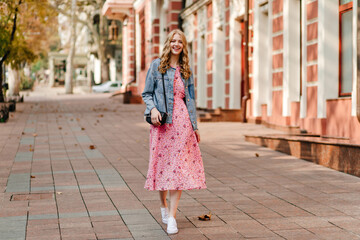 happy curly blonde walks down the street in a denim jacket and a light pink dress on a blurred background of the building