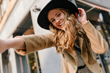 Fototapeta na wymiar Smiling, perky curly young lady takes a selfie on the street. Portrait of a blonde with a gentle make-up in a hat