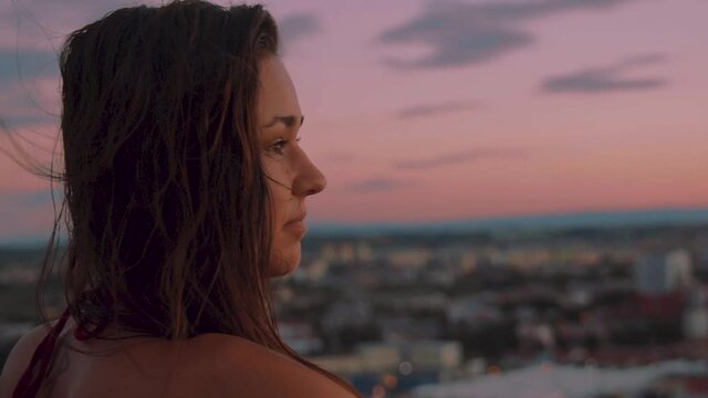 Young brunette woman enjoys peaceful sunset view over skyline of city, pan left