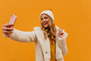 Cheerful, beautiful, energetic girl uses her pink iPhone for selfie. Curly blonde in warm eco fur coat and yellow sweater on orange background.