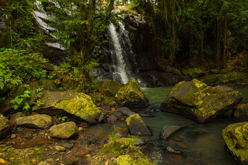 Naklejka na ściany i meble Waterfall landscape. Beautiful hidden waterfall in tropical rainforest. Foreground with big stones. Fast shutter speed. Sing Sing Angin waterfall, Bali, Indonesia