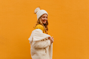 Beautiful curly blonde in orange sweater puts on eco fur coat and looks down flirty - obrazy, fototapety, plakaty