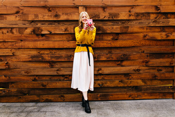 Full-length photo of girl in yellow outfit with black belt. Blonde makes photo on pink camera