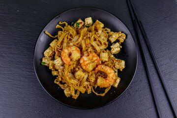 Stir-fried rice noodles Pad Thai with prawns and tofu in a plate on black slate background. Top view. Thai food