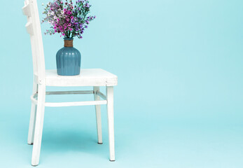 Fototapeta premium Bouquet of artificial lavender bouquet flowers in glass vase on white chair and blue background. Minimal style interior. Copy space.