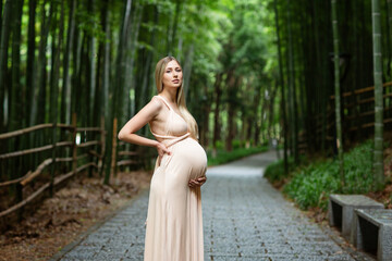 Pregnant woman in evening dress holds hands on belly outdoor in bamboo forest. Pregnancy,...