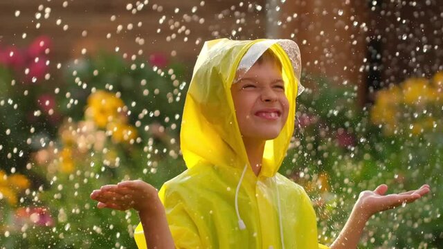 Little happy cute smiling boy child in yellow raincoat and enjoying having fun rainfall. Kid playing with drops rain in sunlight. Happy family summer autumn fall childhood dream concept. 120 slow-mo