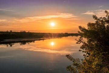 Obraz premium Scenic view at beautiful summer river sunset with reflection on water with green bushes, grass, golden sun rays, calm water ,deep blue cloudy sky and glow on a background, spring evening landscape