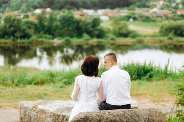 Newlyweds sitting on a stone on the background of the lake in cloudy weather. Rear view.