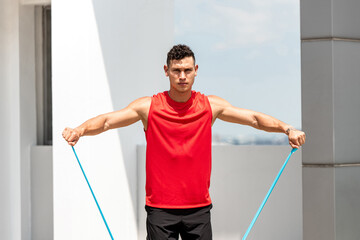 Handsome sports man doing shoulder lateral raise exercise with resistance band outdoors on rooftop...