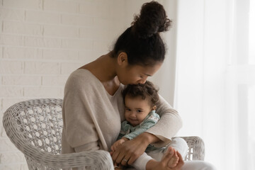 Loving caring young mixed race woman kissing little adorable baby kid, relaxing on comfortable chair. Affectionate african mommy showing devotion to cute boy or girl, spending daycare time together.