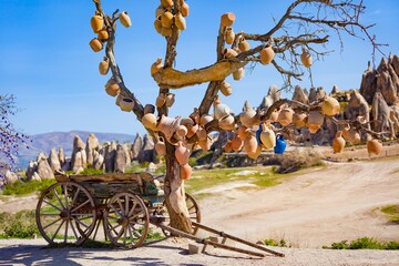 Goreme National Park, Cappadocia, Turkey. Old wooden cart stands under tree with clay pots hanging from its branches. Unusual landscape with conical rocks is in background.