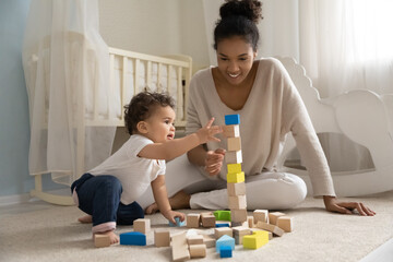 Happy young african american mother or nanny sitting on floor carpet, constructing building with wooden cubes with curious adorable small toddler baby boy girl in children room, childcare concept. - obrazy, fototapety, plakaty