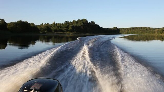 Motor boat with an powerful outboard motor fast gliding on river calm water, beautiful view from stern on transom Wake at Sunny summer day on trees on shore and blue sky background, boating lifestyle