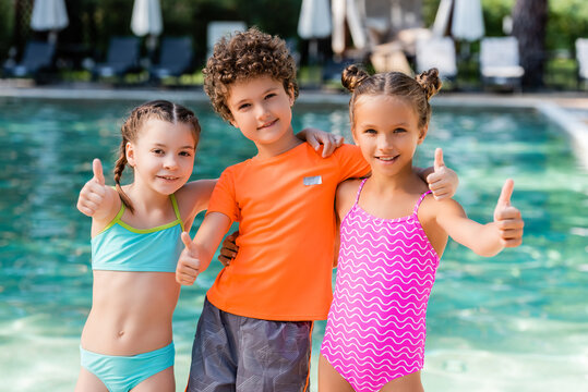 curly boy in t-shirt and girls in swimsuits showing thumbs up near pool