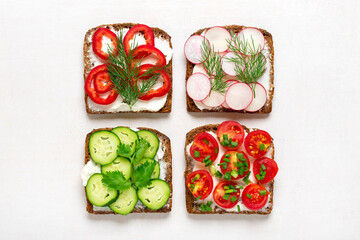 Variety of sandwiches for breakfast - slice of whole grain dark bread, pepper, cream cheese, cucumbers, radishes, cherry tomatoes, garnished with dill, green onions on white table Top view Flat lay