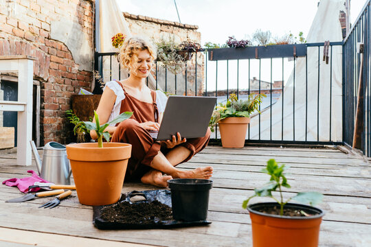 Blond woman gardener wear brown overalls, sitting on wooden floor in terrace resting, using laptop after work, smiling and speaking on video call surrounded by plants. Home gardening concept.