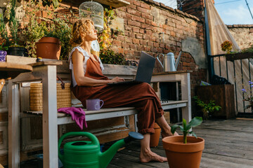 Searching for the best advice. Romantic blond haired smiling female gardener in overalls surfing the internet on her laptop sitting near her plants pots, equipment, watering can in terrace outdoor.