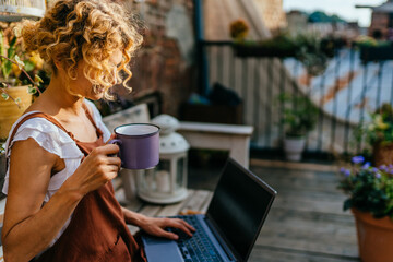 Blond woman gardener wears brown overalls relaxing after work in terrace focusing on cup of tea and working with laptop.