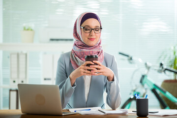 Beautiful young working woman in hijab, suit and eyeglasses sitting in office, smiling. Portrait of confident muslim businesswoman drinking coffee. Modern office with big window, 