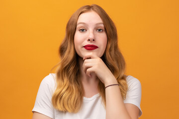 Photo of Pensive young woman with wavy redhead standing with hand raised on chin and looking thoughtfully into a camera