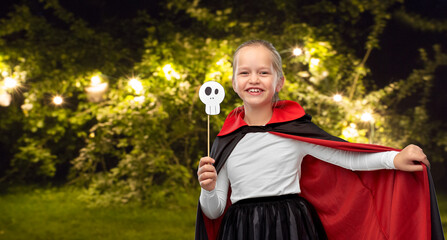 halloween, holiday and childhood concept - girl in dracula costume with black cape holding scull...