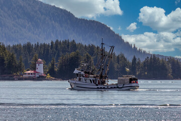 Shot of a commercial fishing boat sailing in a harbour in Sitka, AK. Green forest, mountains, beautiful blue sky with clouds, and a lighthouse in the background.