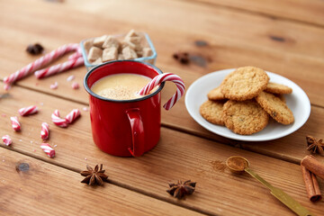 christmas and seasonal drinks concept - red cup of eggnog with candy cane, oatmeal cookies, star anise and cinnamon on wooden background