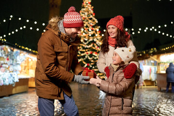 family, winter holidays and celebration concept - happy mother, father and little daughter with gift at christmas market on town hall square in tallinn, estonia