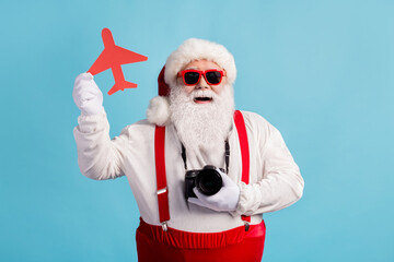 Photo of retired old man grey beard hold paper plane figure camera tourism check-in registration low-cost wear santa x-mas red costume suspender sunglass cap isolated blue color background