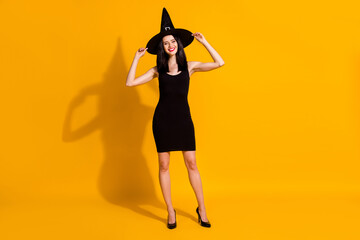 Full body size photo of cute pretty young witch lady hands cap smiling preparation october theme masquerade wear black headwear high heels dress isolated bright yellow color background