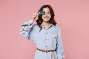 Smiling young brunette woman 20s wearing casual blue shirt dress posing holding in hand covering eye with credit bank card looking camera isolated on pastel pink colour background, studio portrait.