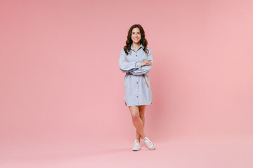 Full length portrait of smiling funny young brunette woman 20s wearing casual blue shirt dress posing standing holding hands crossed looking camera isolated on pastel pink colour background in studio.